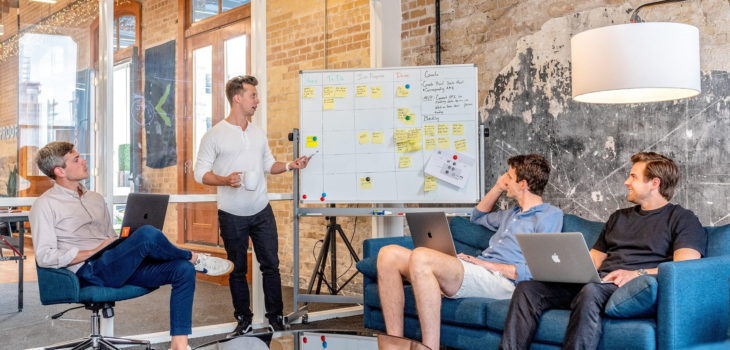 Three middle aged men listen to a male colleague present at a whiteboard