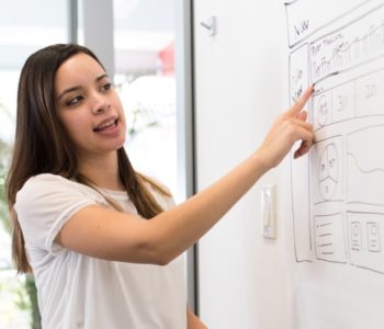 brown haired woman in a white shirt presents ideas while pointing at a flip chart at work