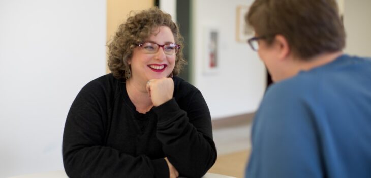 Curly-haired, plus-sized woman manager with glasses and red lipstick smiles and listens intently to her male direct report
