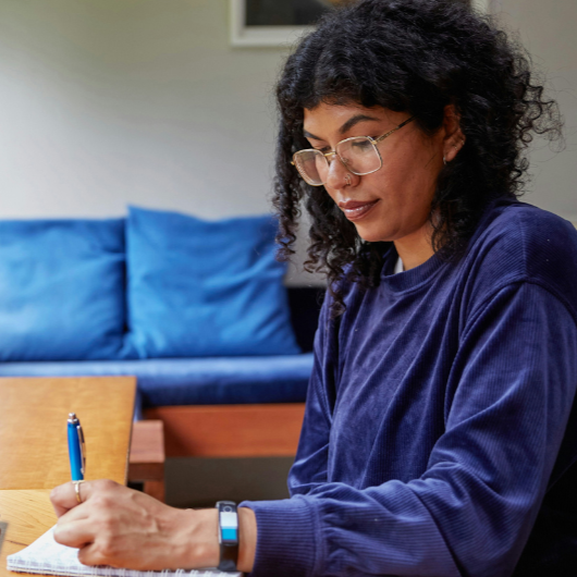 Black woman with long natural curly hair and wire-rimmed glasses in an indigo corduroy sweatshirt prepares for a job search on her laptop and takes notes in her journal