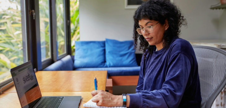 Black woman with long natural curly hair and wire-rimmed glasses in an indigo corduroy sweatshirt prepares for a job search on her laptop and takes notes in her journal sitting in a natural-light filled room with large windows and green flax bushes outside