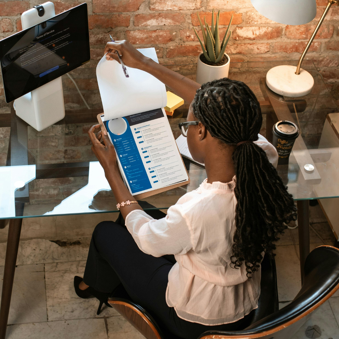 A woman of colour wearing a pale pink blouse, black pants and black high heels, with long braids, sitting at glass desk in an office reviewing a resume on a clipboard