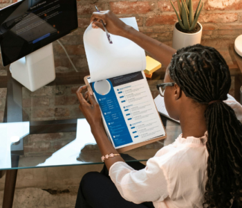 A woman of colour wearing a pale pink blouse, black pants and black high heels, with long braids, sitting at glass desk in an office reviewing a resume on a clipboard