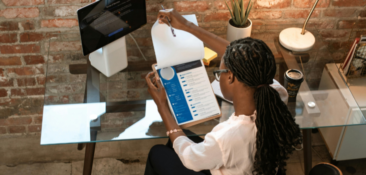 A woman of colour wearing a pale pink blouse, black pants and black high heels, with long braids, sitting at glass desk in an office reviewing a resume on a clipboard