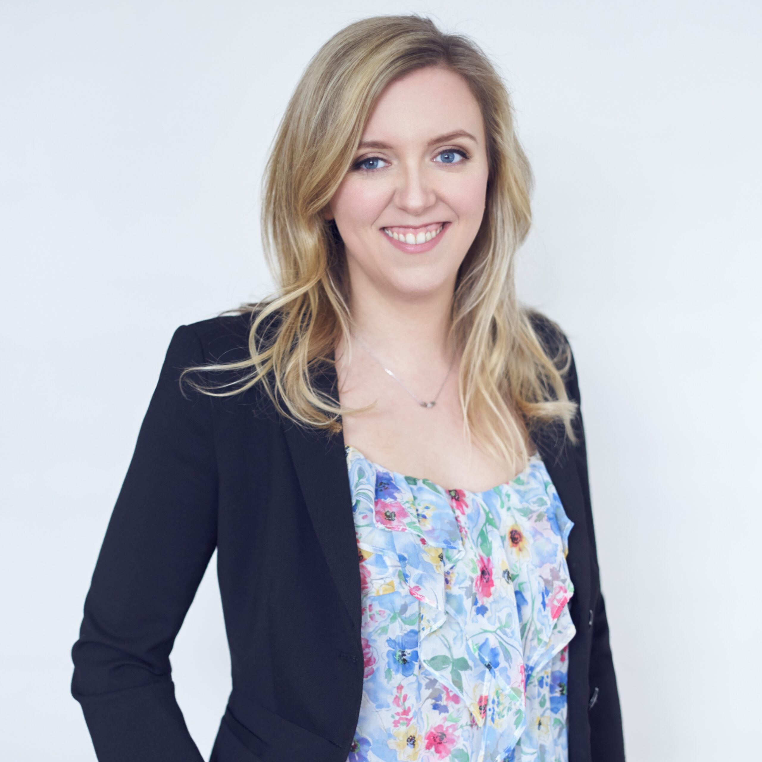 Holly Burton, a white woman, with blonde hair wearing a navy-blue blazer and floral blouse, smiles while standing facing the viewer.