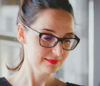 A Caucasian woman wearing a black jumper, tortoiseshell rimmed glasses and bright red lipstick with hair pulled back in a ponytail, looking down at a computer screen