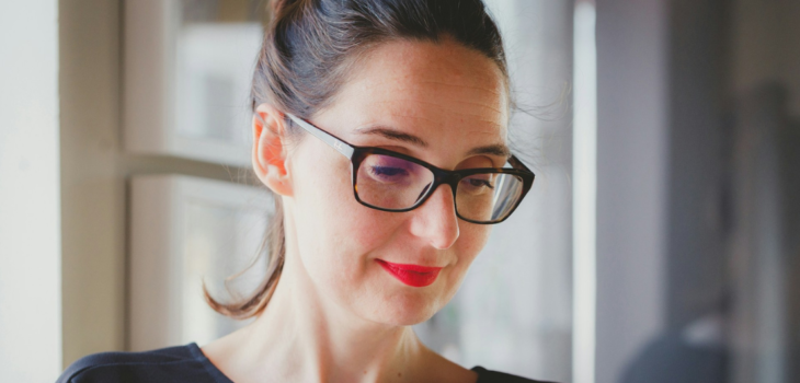 A Caucasian woman wearing a black jumper, tortoiseshell rimmed glasses and bright red lipstick with hair pulled back in a ponytail, looking down at a computer screen