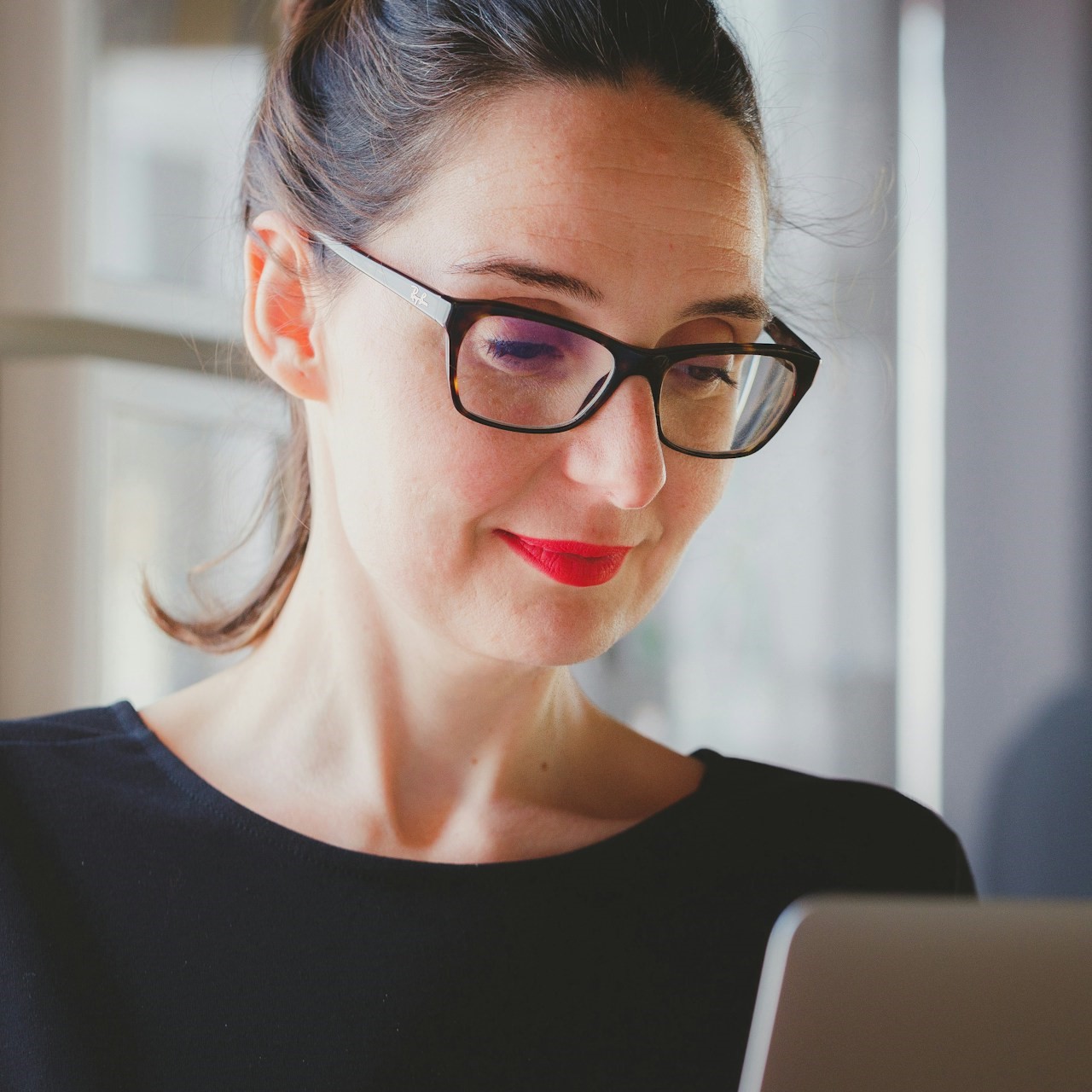 A Caucasian woman wearing a black jumper, tortoiseshell rimmed glasses and bright red lipstick with hair pulled back in a ponytail, looking down at a computer screen